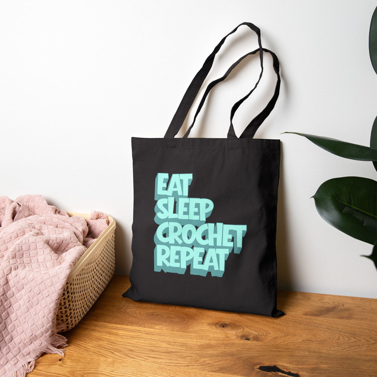 Black tote bag with 'Eat Sleep Crochet Repeat' text on a wooden surface with a plant in the background