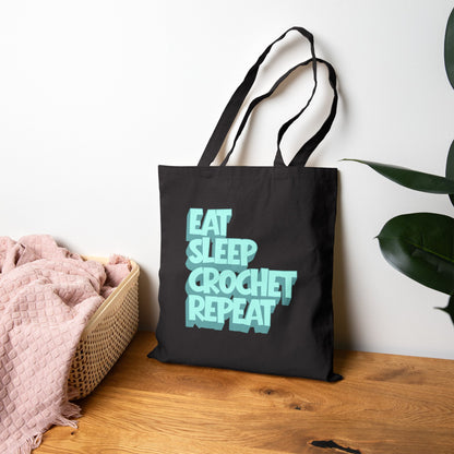 Black tote bag with 'Eat Sleep Crochet Repeat' text on a wooden surface with a plant in the background