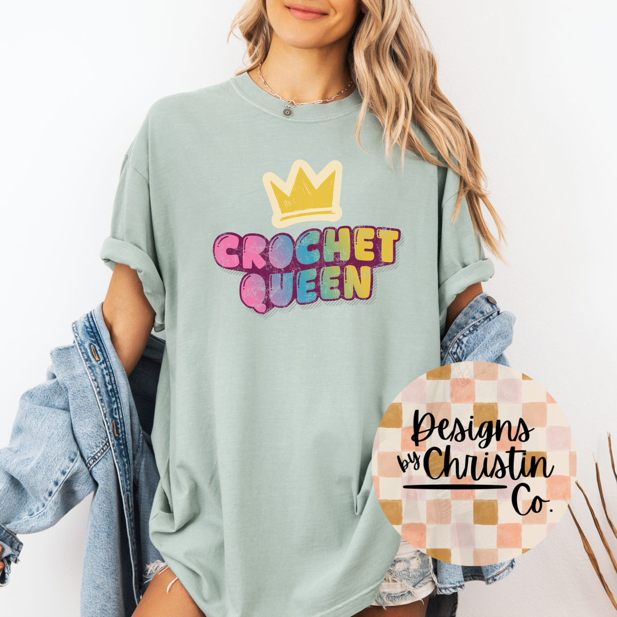 Person wearing a light green t-shirt with 'Crochet Queen' text and a crown graphic, holding a circular sign with 'Designs by Christin Co.'.