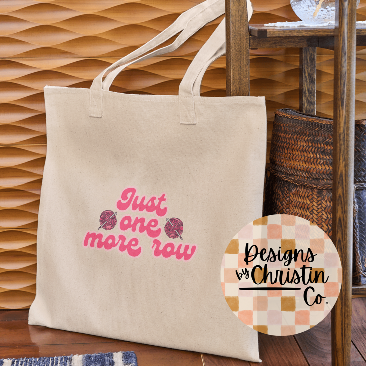 Beige tote bag with pink text 'Just one more row' on a wooden surface, with a circular logo in the corner.