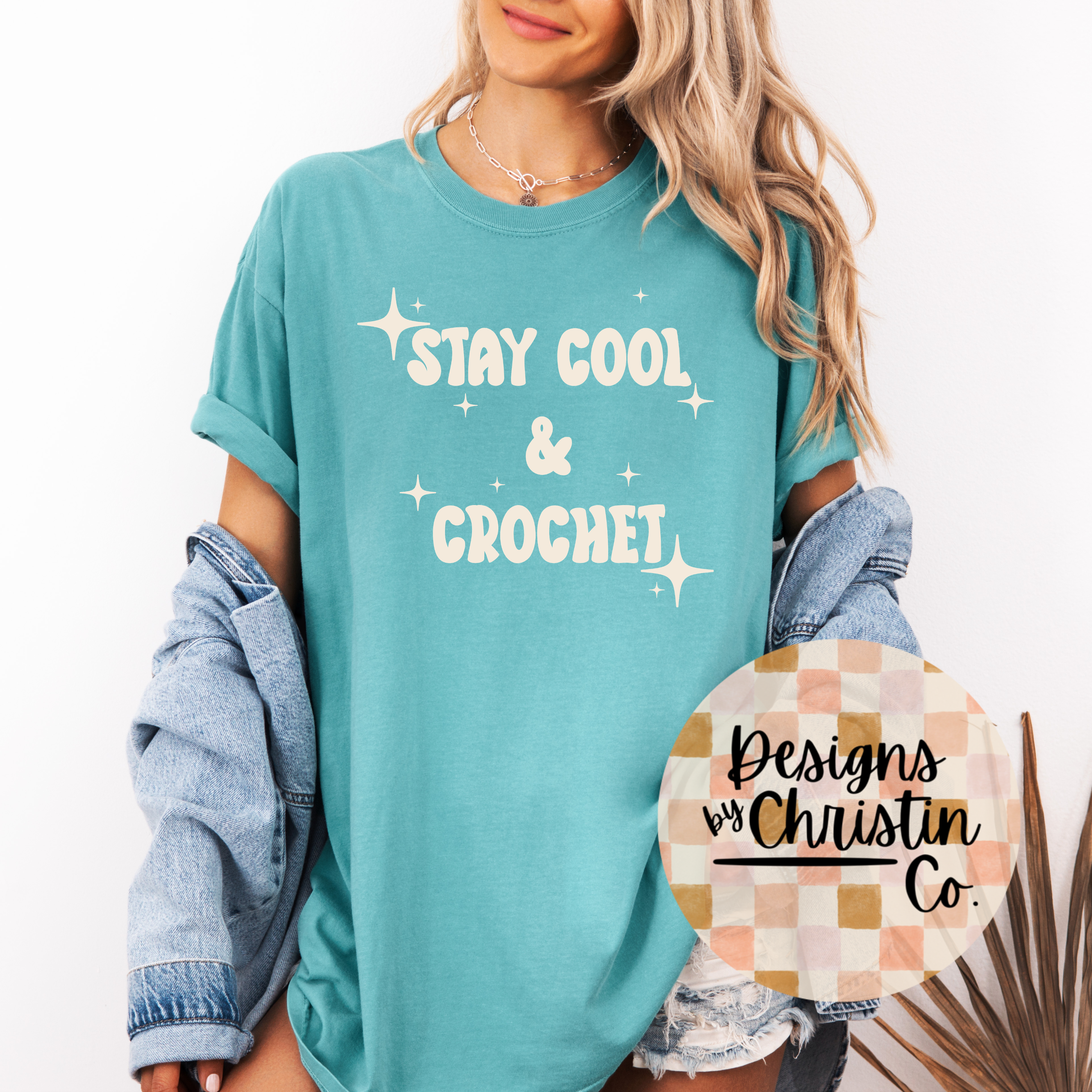 Person wearing a teal t-shirt with 'Stay Cool & Crochet' text, holding a circular sign with 'Designs by Christin Co'.